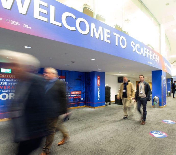 Image of a corridor with people walking down it. Blue and red branded walls display the words Welcome to Scaffex24.