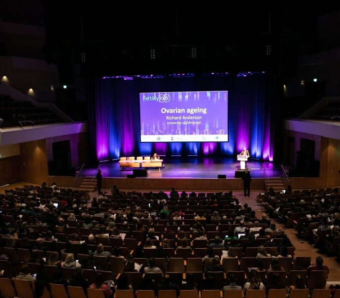 An auditorium is full of hundreds of people sitting facing a stage. On stage a presentation is displayed with the title 'Ovarian Ageing.' The presentation is being delivered by a person standing at a podium on the right hand side of the stage.