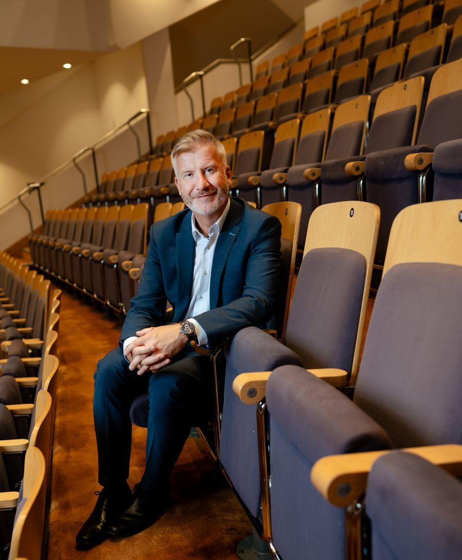 Man in his fifties in a navy suit and blue shirt sitting in an auditorium with his hands together looking at the camera. Behind him are rows of empty theatre seats.