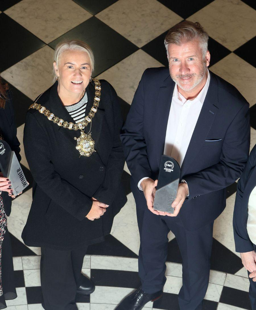 Four people stand on a black-and-white checkered floor, smiling at the camera. Two of them hold awards labelled “C&IT Awards 2025 Winner.” The person standing in the centre is wearing a Lord Mayor ceremonial chain. All are dressed in formal business attire.