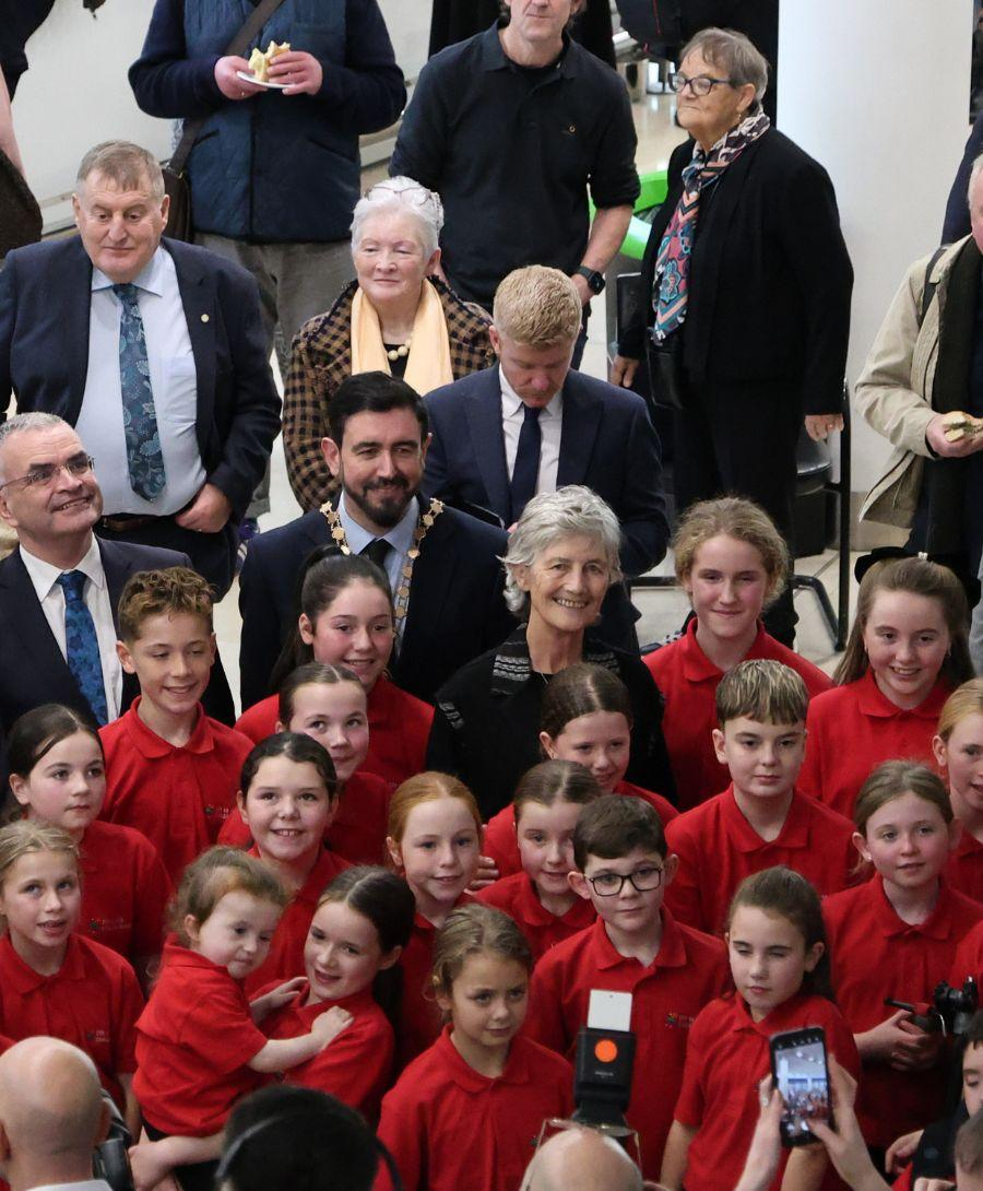 Group photo showing smiling school children in red uniforms surrounding a woman in the center, with three men standing to the side and others watching in the background.