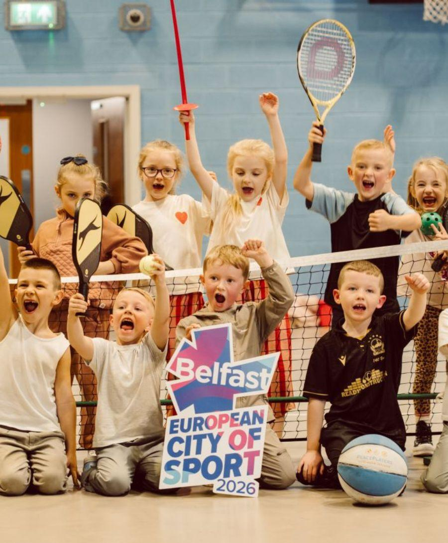 Image of children on a tennis court indoors cheering for press shot. Some are holding tennis rackets as props meanwhile one in the middle is holding a prop to mark the achievement
