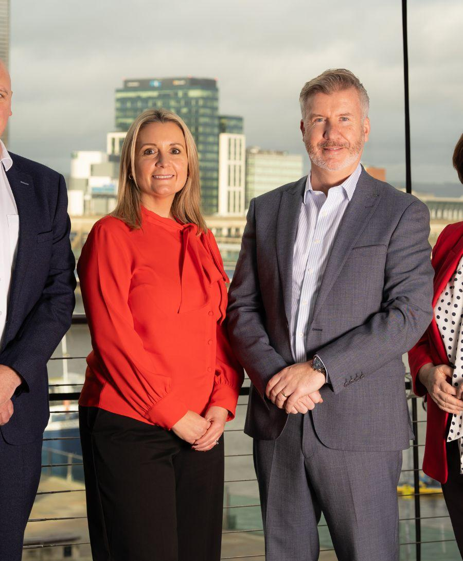 Two men and two women smile for press shot against window with Belfast city skyline in the background