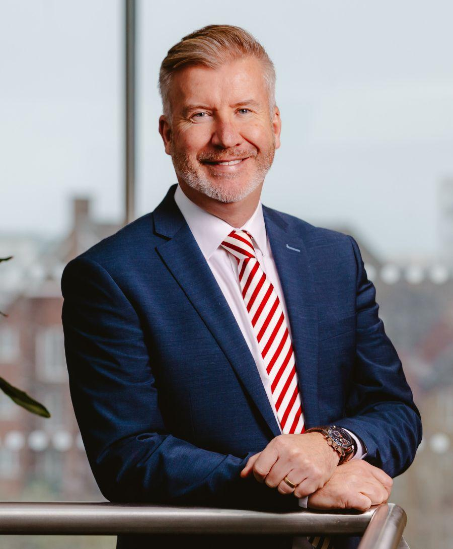 Image of man smiling for headshot with plant beside him and window in the background