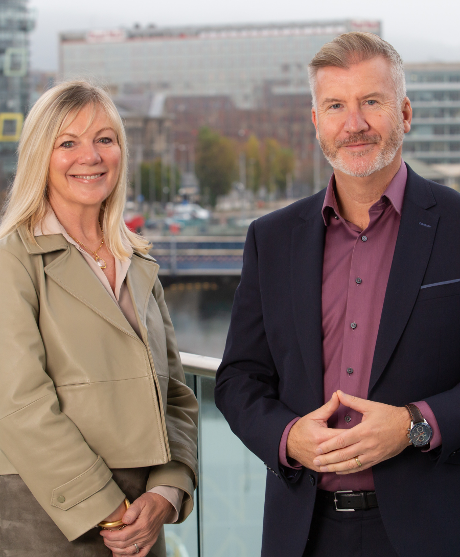 Two people standing outdoors on a balcony overlooking a river and cityscape. The woman on the left is wearing a light beige jacket and smiling, while the man on the right is wearing a dark suit with a maroon shirt and also smiling. Buildings and a bridge are visible in the background.