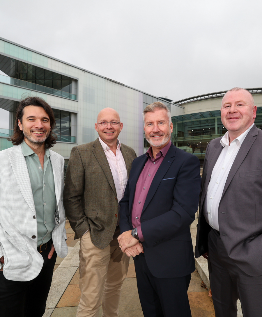 Four men stand in a line smiling at the camera wearing professional business attire. Behind them is a large glass fronted building with protruding balconies.