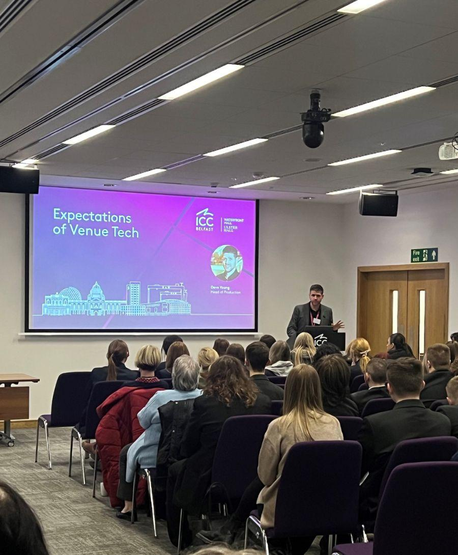 ICC Belfast Head of Production Dave Young addresses attendees at the Future You 'on the road' event in Meeting Room 1. Image shows Dave at the top right in front of a screen looking down at number of people sitting on chairs facing forward.