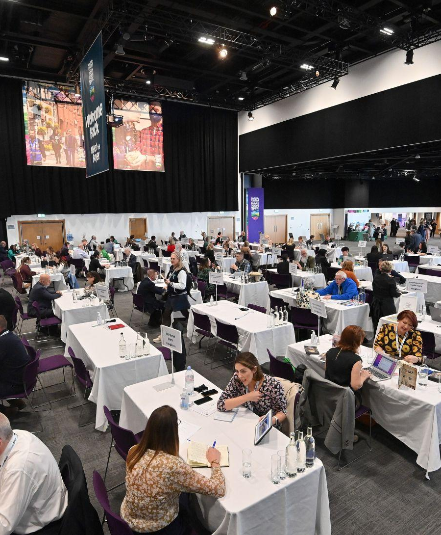 Delegates seated in Hall 1 at rectangle tables of four with a large banner at the right side of the image that reads 'welcome back, meet the buyer'
