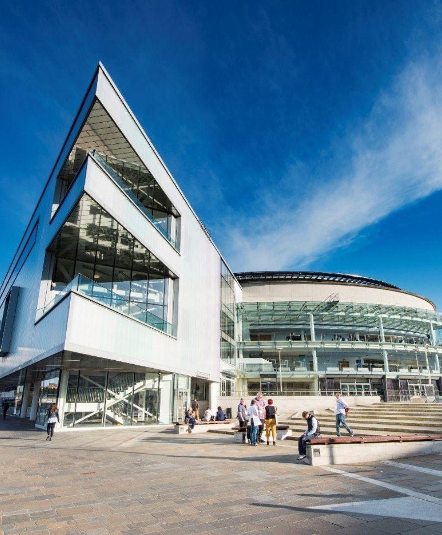 Image of the exterior of ICC Belfast taken in the sunshine in front of the entrance beside the river with individuals on the steps outside the building.