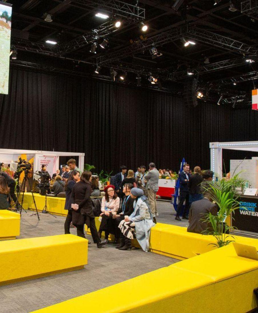 Men and women sitting in the exhibition space with long yellow seating, greenery and digital screens.