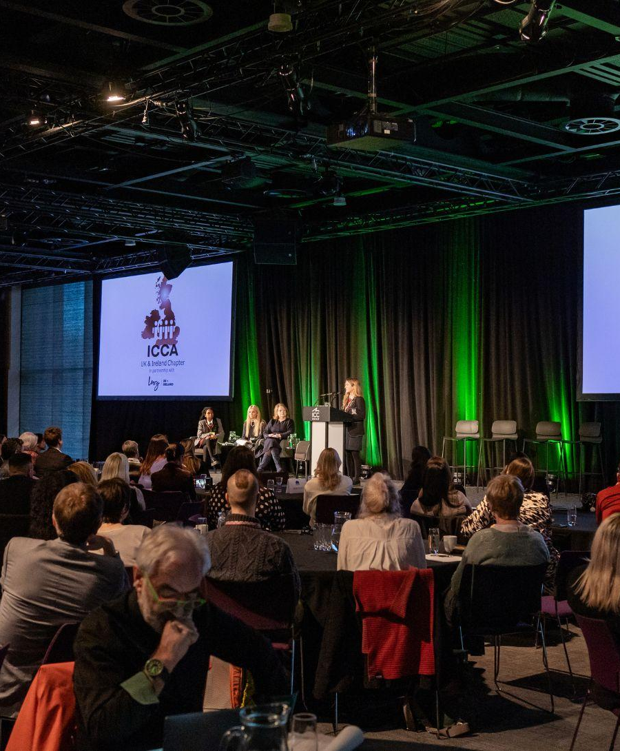 The audience is paying attention to a SistersIN pupil who is giving a speech on stage, there are also digital screens in the background with the ICCA logo and green lights displayed.