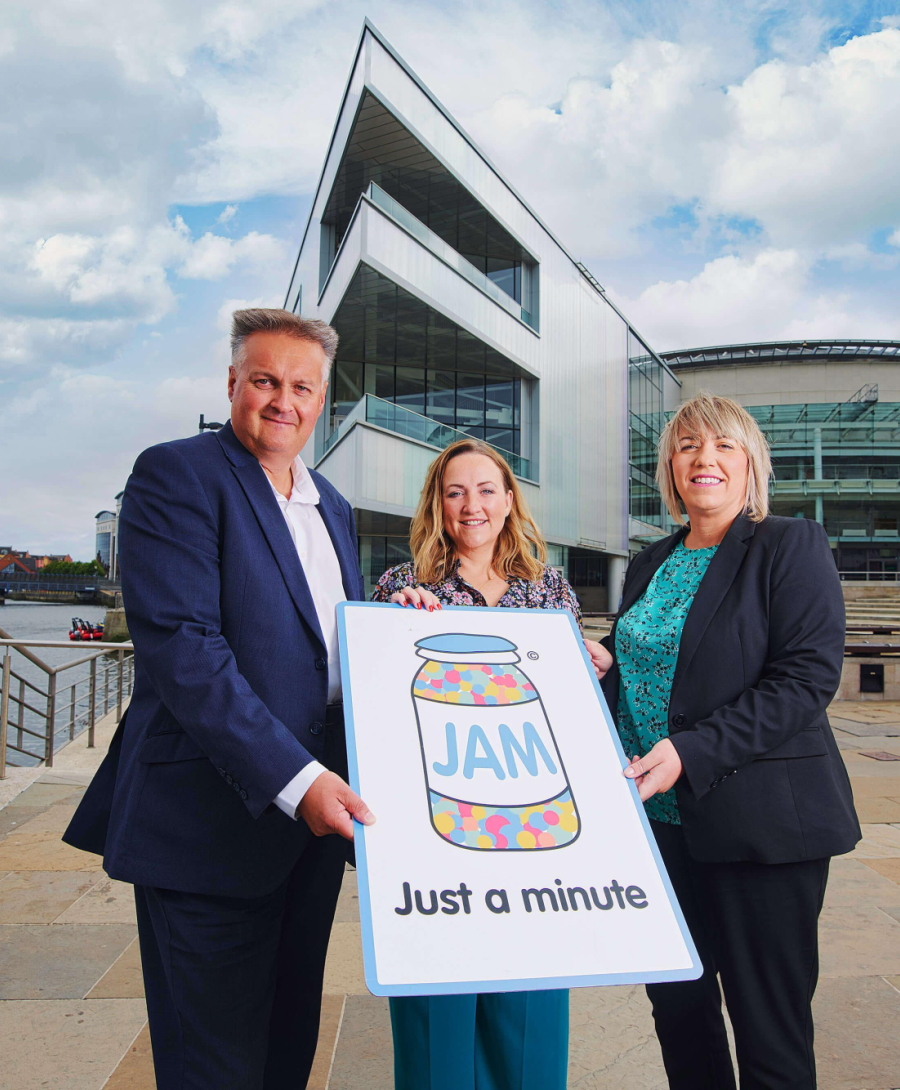 Image shows three people holding large cardboard cut out in from of ICC Belfast building which has JAM card logo and the words 'Just a minute'.