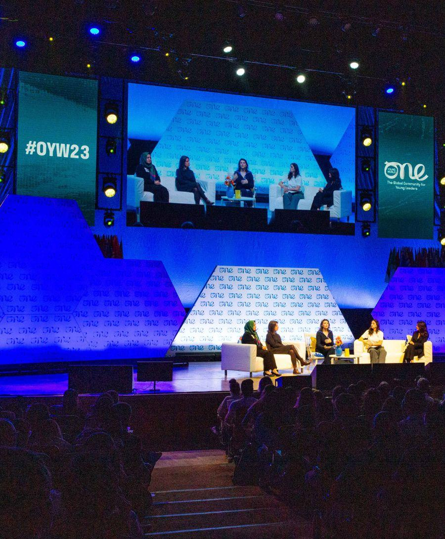 One Young World Summit 2023 in progress in the Main Auditorium with a panel discussion on going and delegates seated.