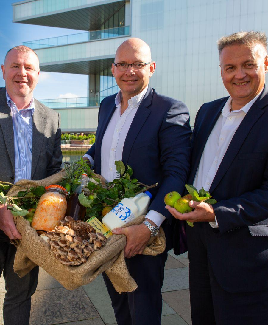 Three men stand in front of building with a woven basket filled with food produce including milk, apples, mushrooms and lettuce
