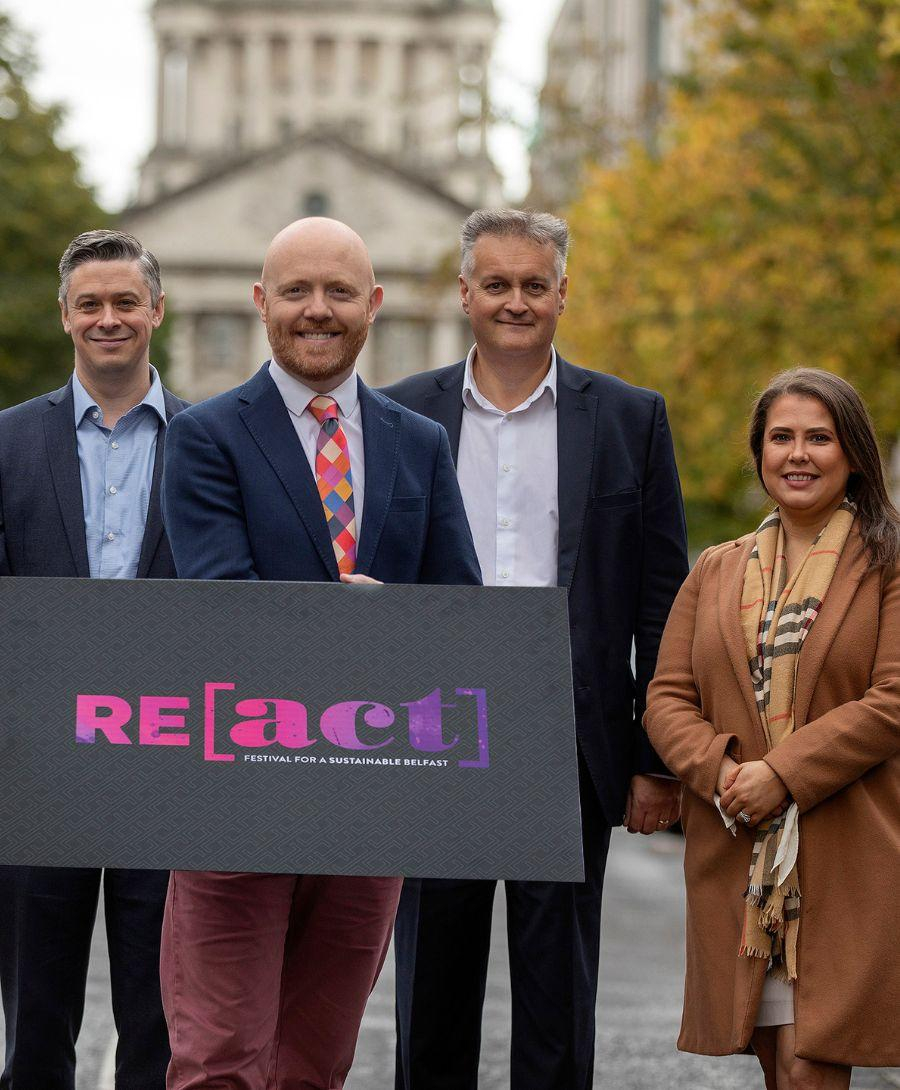 Three men and one woman stand on street surrounded by trees holding black sign with the letters REACT
