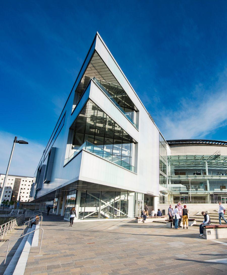 White pointed building which shows three levels, two with a balcony and to the side is a curved building with windows. Multiple adults standing and sitting outside the building in the sunshine.