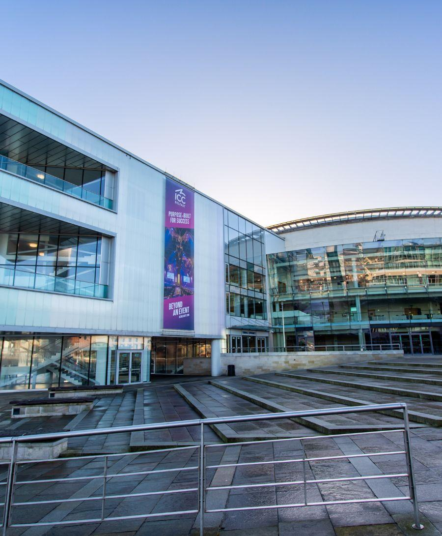Image is a view of the exterior of ICC Belfast showing the Riverside entrance and the bars.
