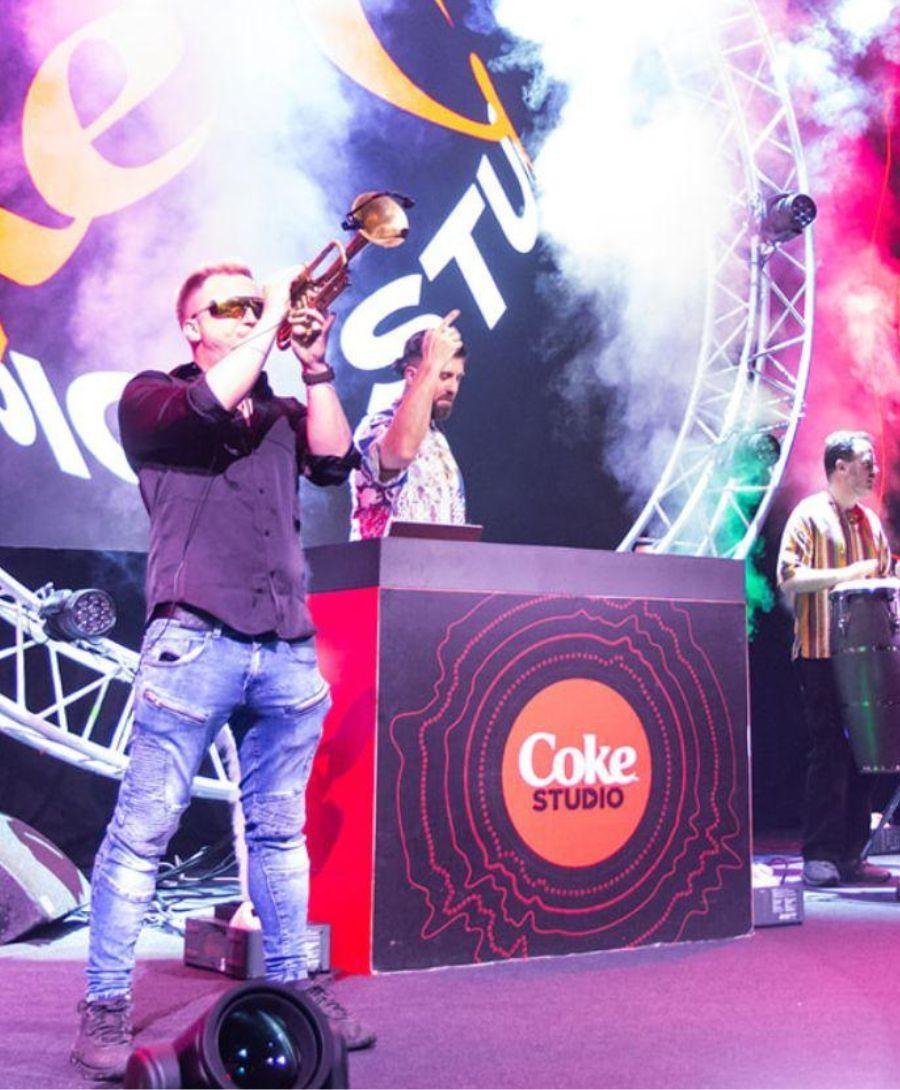 Man playing a trumpet on a stage in Hall 1 with a DJ playing in front of decks branded Coca-Cola. Digital signage also shows the words 'Coke Studio'