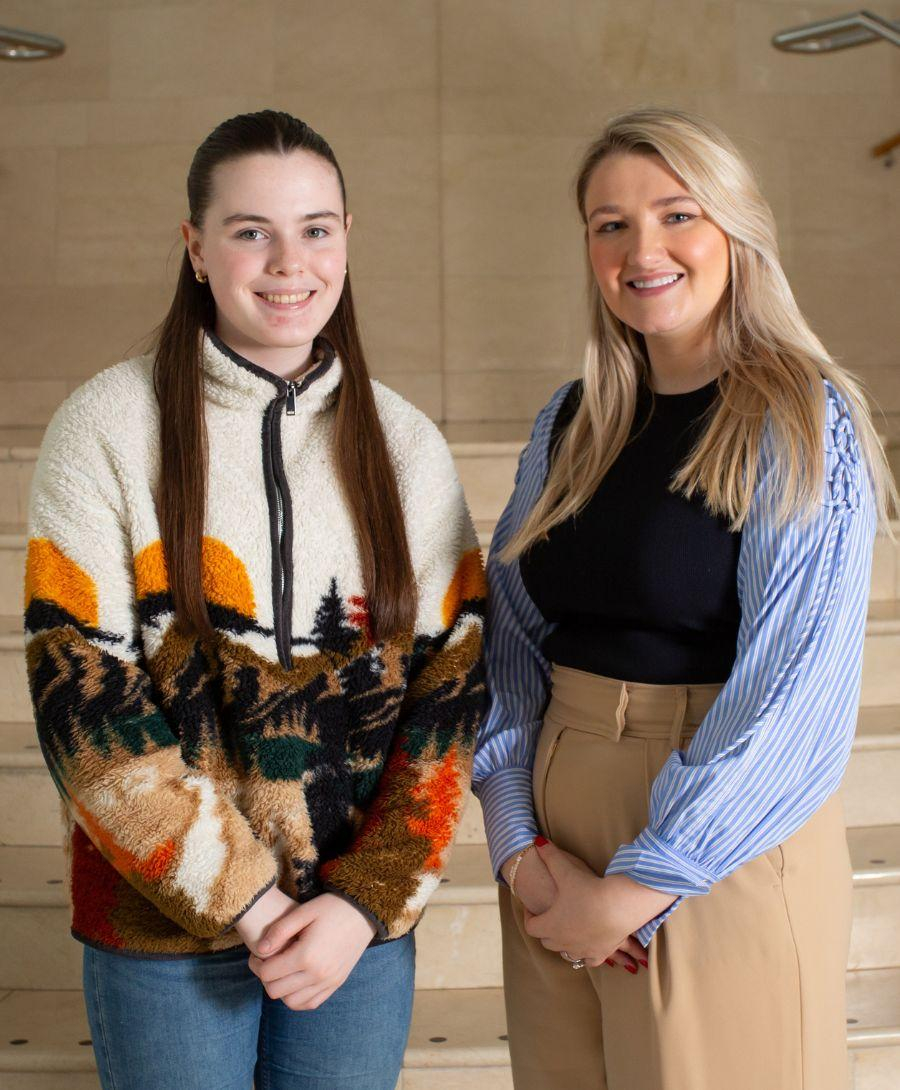 Two females stand in front of a cream marble staircase. One girl to the left has long brown hair and is wearing a white fleece with brown, mustard and black colours faded from the bottom. The girl on the right had shorter blonde hair and is wearing a blue shirt with beige trousers.