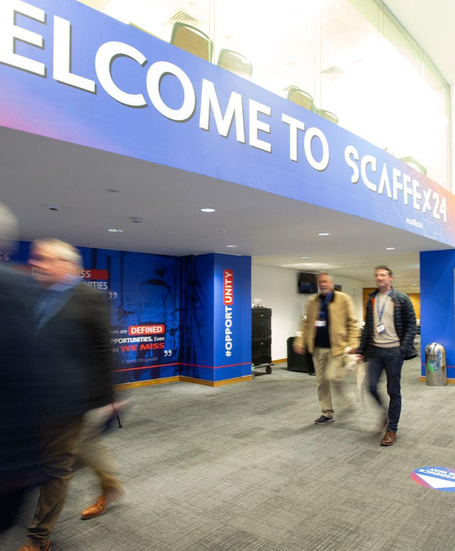 Image of a corridor with people walking down it. Blue and red branded walls display the words Welcome to Scaffex24.
