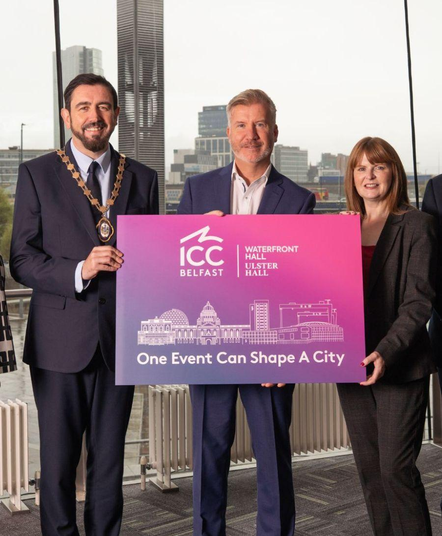 Two men in suits and a lady in a suit stand in front of window holding a pink sign with the words 'One Event Can Shape A City'