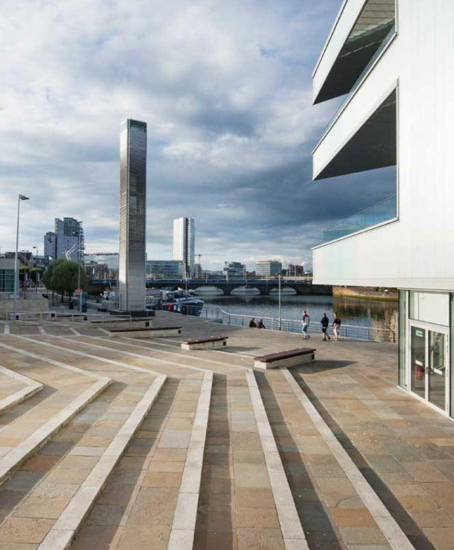 This image shows a small part of the exterior of ICC Belfast with steps and benches to the side. The River Lagan, Queen's Bridge Belfast and Obel Tower are visible in the background.