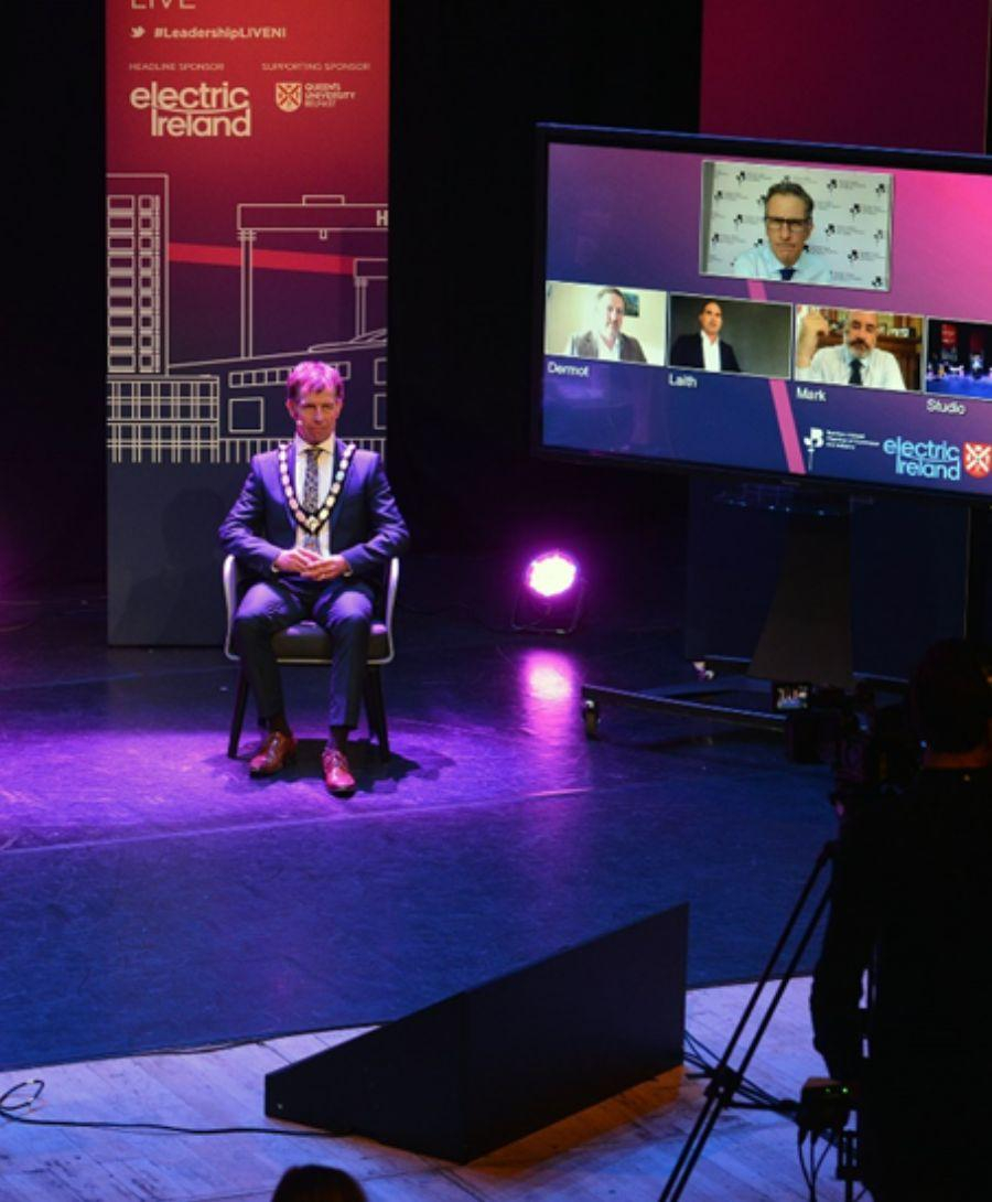 Three people seated on a stage for a discussion. Banners are displayed at either side of the people involved and two the right of them is a large screen with other members on a video call to join the hybrid event.