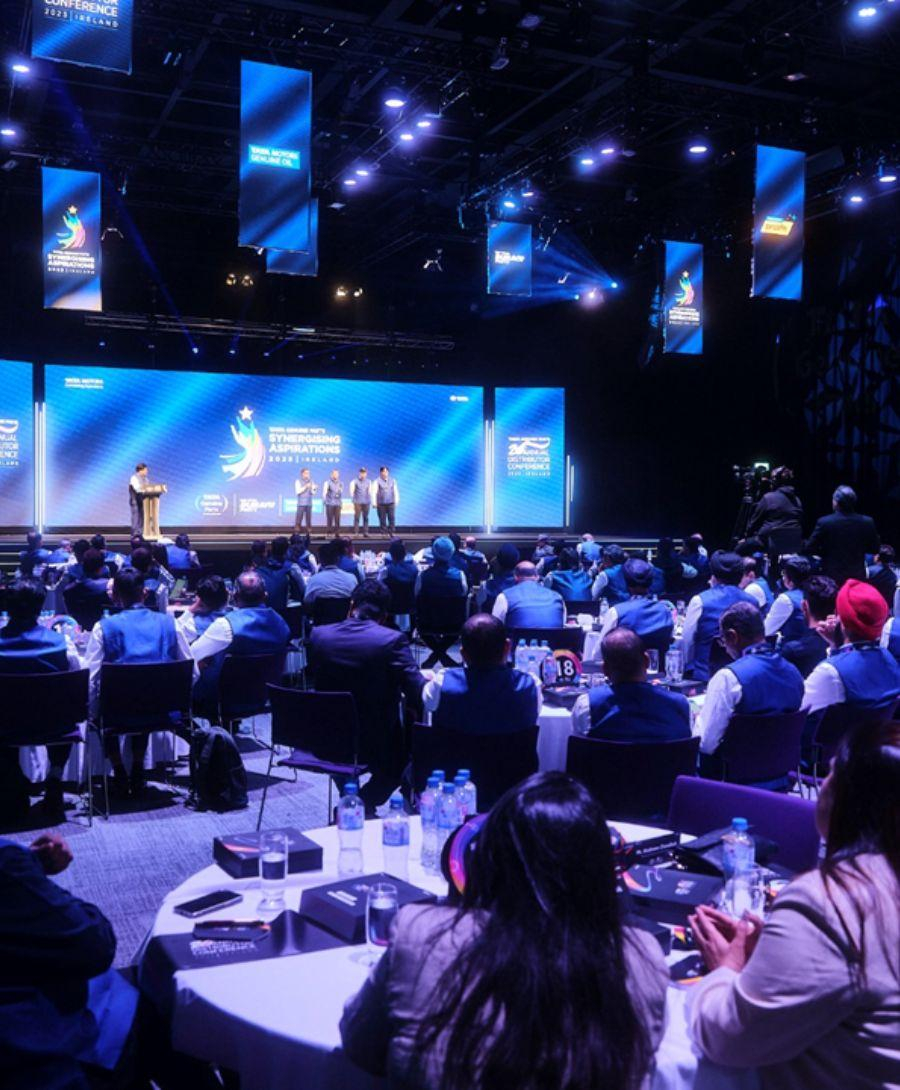 Delegates seated at round tables for the Tata Genuine Parts for their Annual Distributor Conference. They are facing a stage with four members stood to the right and one at a podium on the left. The large screen behind them reads, 'Tata Genuine Parts, Synergising Aspirations, 2023, Ireland.'