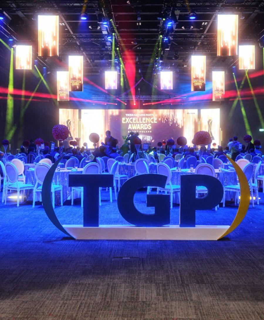 Hall 1 set up for the TATA Genuine Parts (TGP) Annual distributor conference award ceremony, large branding statue that reads (TGP) with round tables set up in the back facing a stage with coloured lights and a large digital screen that reads TATA Genuine Parts Excellence awards.
