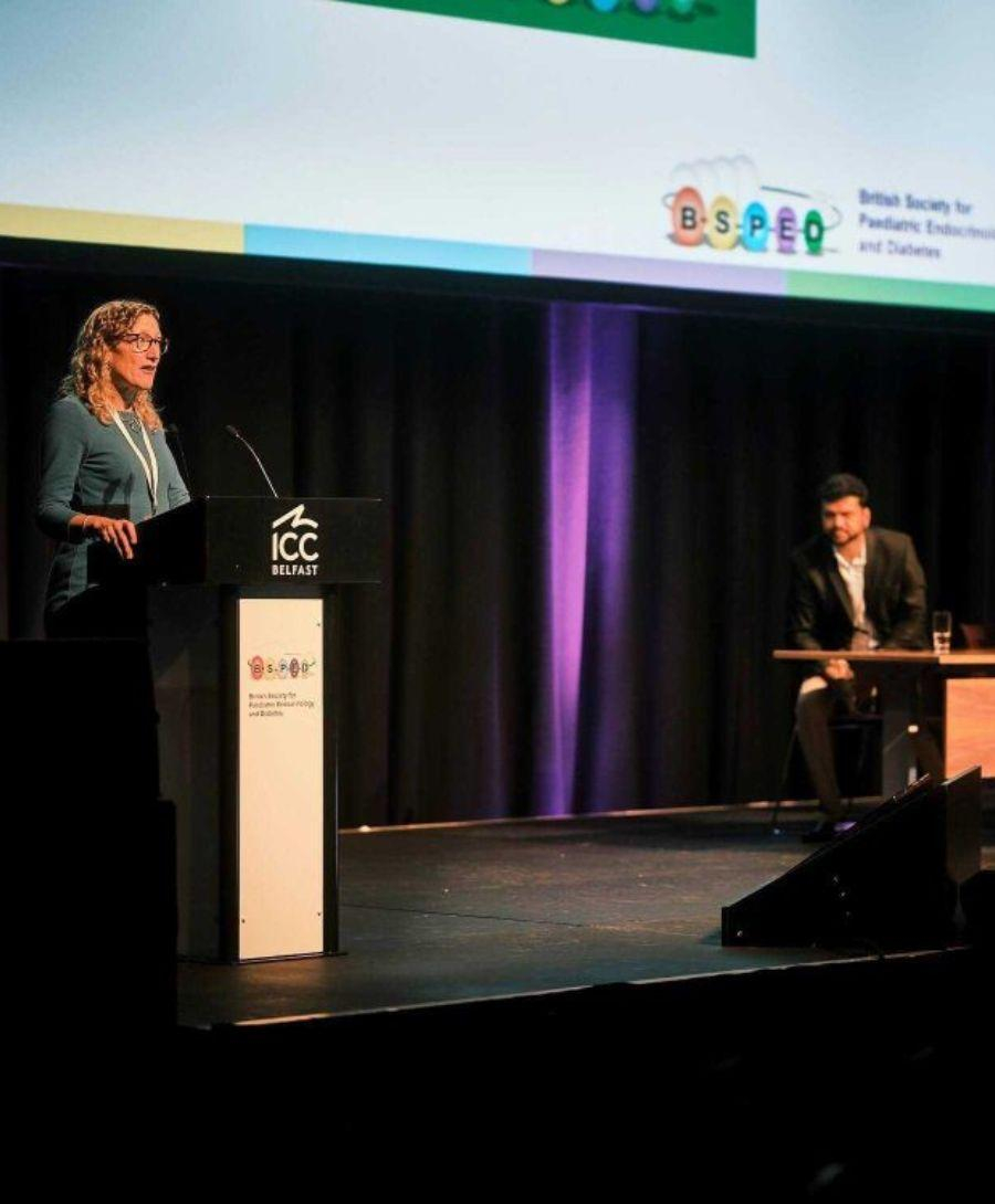 Woman stood at a podium speaking in to a microphone, on stage at the British Society for Paediatric Endocrinology and Diabetes event. To the right is a man sat on the panel listening.