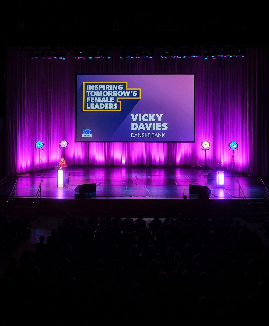 Main Auditorium set up for the SistersIN celebration event. Vicky Davies is stood at a podium delivering a presentation to the attendees and is stood in front of a large digital screen that displays her name and reads, ' Inspiring tomorrow's female leaders'