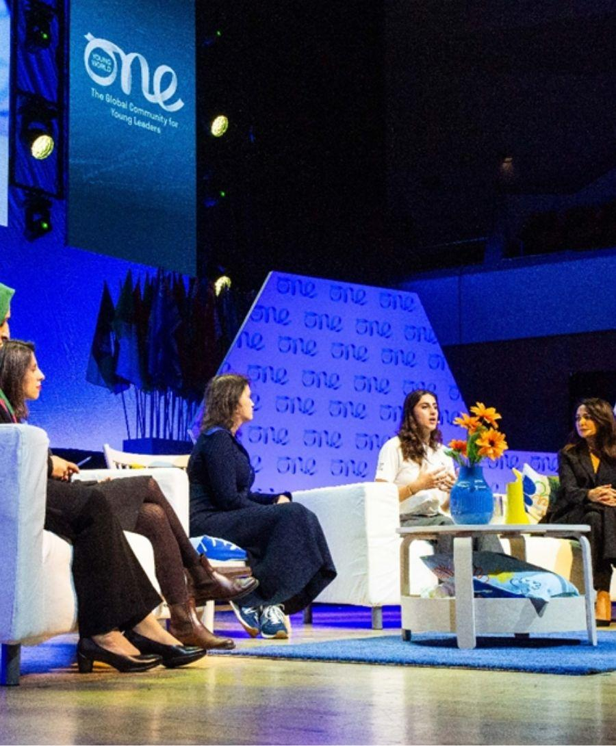Five female delegates seated on the main auditorium stage during the one young word event with the company branding distributed behind them, alongside a large screen that shows close up shots of each delegate.