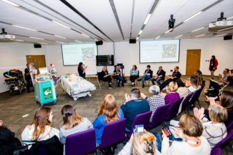 Delegates seat in a semi circle around a hospital bed demonstration as a presenter showcases results on two digital screens position on a wall in a meeting room space at ICC Belfast for the Association of British and Irish Critical Care Nurses joint conference in October 2022
