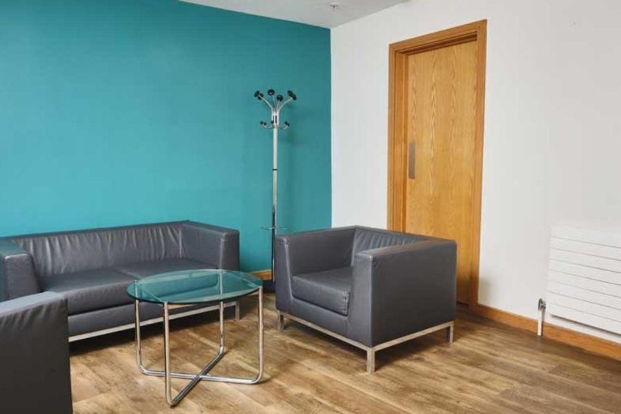 This image shows one corner of the Mezzanine Rooms at ICC Belfast with one blue wall and one white wall with a wooden door. Two grey sofas, a grey armchair and a coffee table are also in the room.