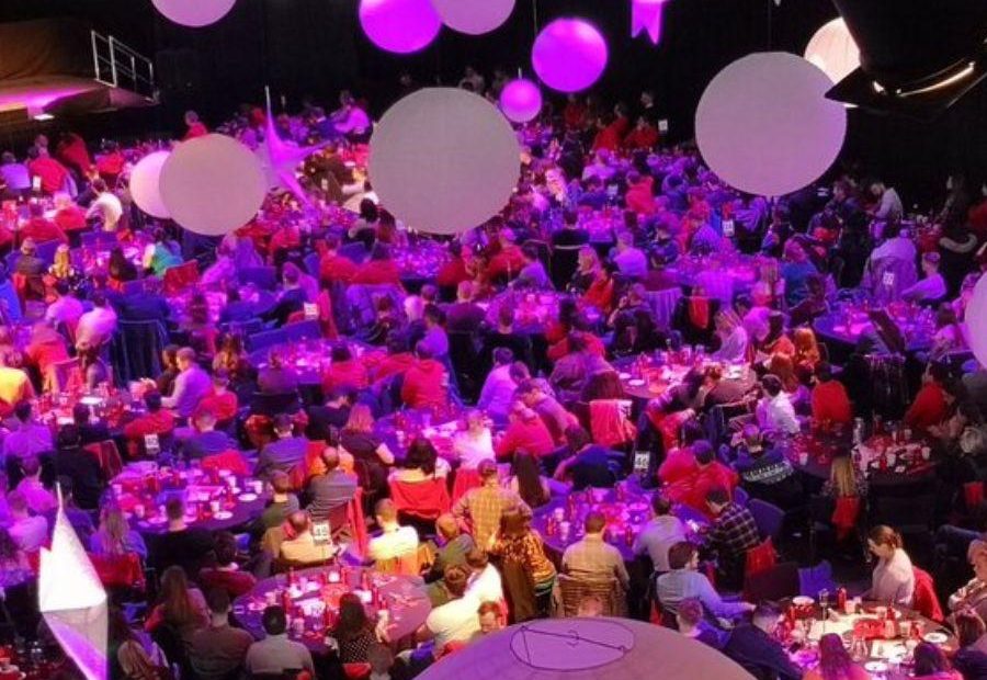 Image shows more than thirty tables full of people at a Christmas party in ICC Belfast. There is a stage in the background and lantern decorations and strings of lights hanging from the ceiling.