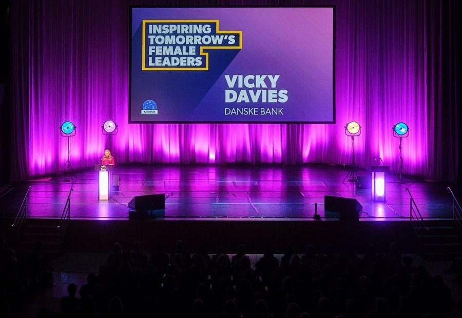 Main Auditorium set up for the SistersIN celebration event. Vicky Davies is stood at a podium delivering a presentation to the attendees and is stood in front of a large digital screen that displays her name and reads, ' Inspiring tomorrow's female leaders'