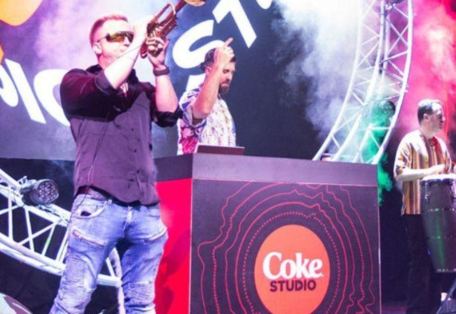 Man playing a trumpet on a stage in Hall 1 with a DJ playing in front of decks branded Coca-Cola. Digital signage also shows the words 'Coke Studio'