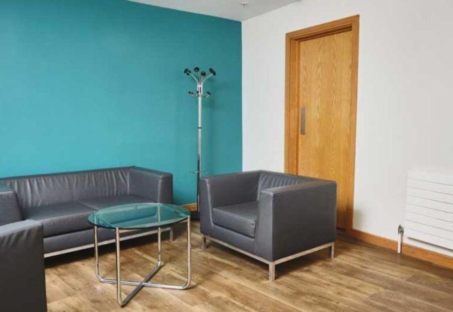 This image shows one corner of the Mezzanine Rooms at ICC Belfast with one blue wall and one white wall with a wooden door. Two grey sofas, a grey armchair and a coffee table are also in the room.