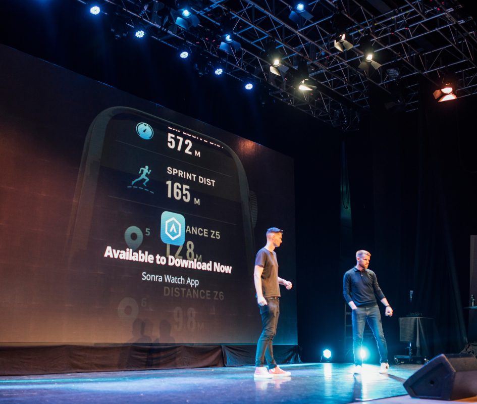 Tech entrepreneurs presenting the Sonra Watch App on stage at ICC Belfast during a live technology conference, with a large digital screen showcasing fitness tracking and app features behind them.