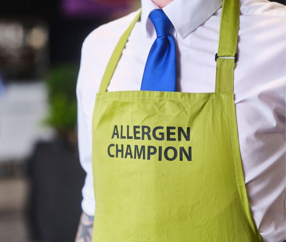 Close up shot of a person wearing a green apron that reads, 'Allergen Champion' all in uppercase, large font.