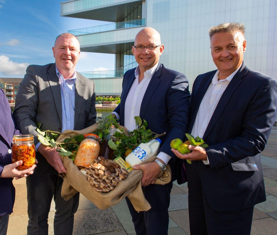 Three men stand in front of building with a woven basket filled with food produce including milk, apples, mushrooms and lettuce