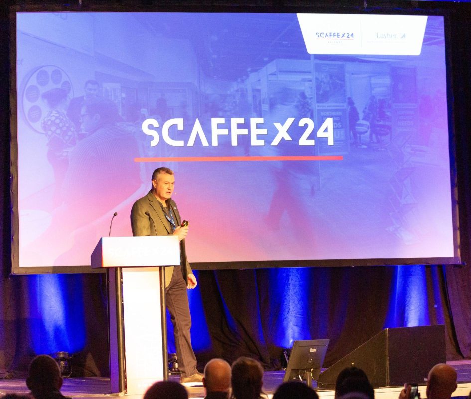 Man stands on stage at lecturn facing a crowd presenting in front on large digital screen with the words 'SCAFFEX24" on display