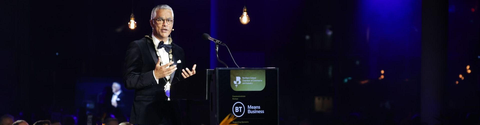 Man in Presidential Chains addresses audience in a gala dinner setting